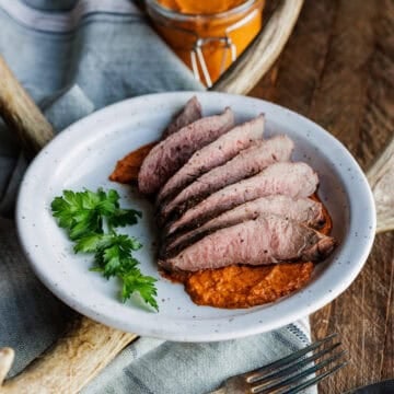 sliced venison steak on plate with romesco sauce and parsley.